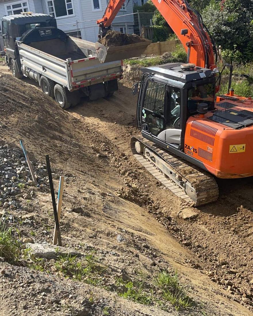 Excavator loading spoil into a tipper truck on a sloping section beside a house
