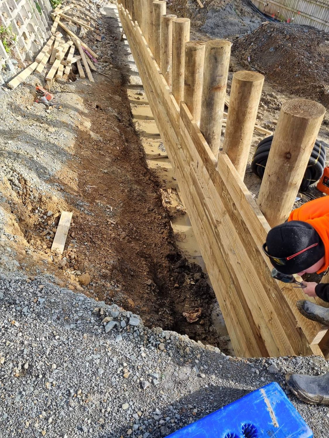 Worker fixing timber lagging behind the poles of a retaining wall, with drainage gravel in the trench
