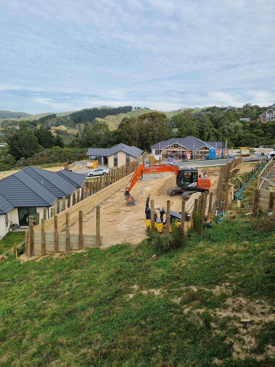 Excavator and roller on building platforms in a hillside subdivision behind a timber pole wall