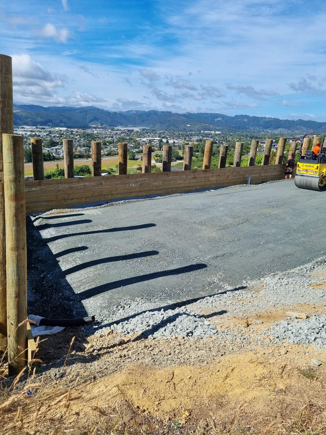 Compacted metal pad behind a new timber pole retaining wall overlooking a town