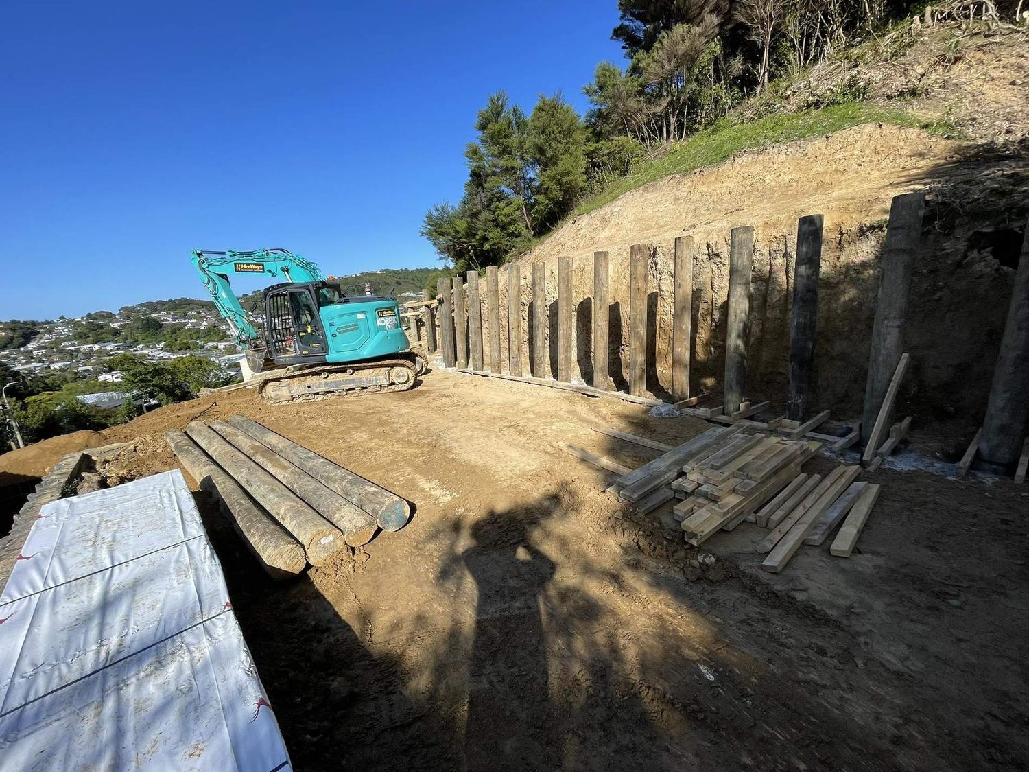 Excavator parked by a timber pole retaining wall under construction, with timber stacked in front