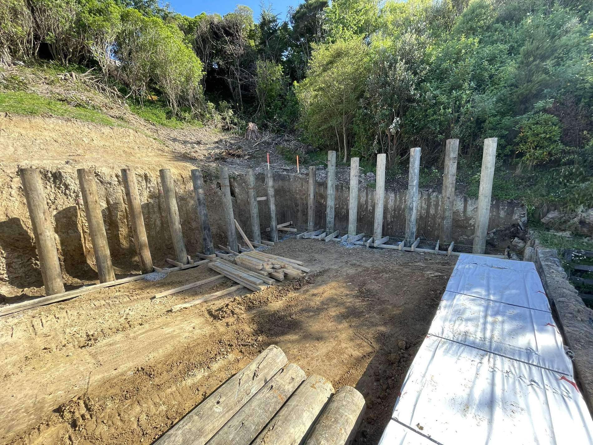 Timber pole retaining wall under construction at the foot of a bush-covered bank, with lagging boards stacked on the pad