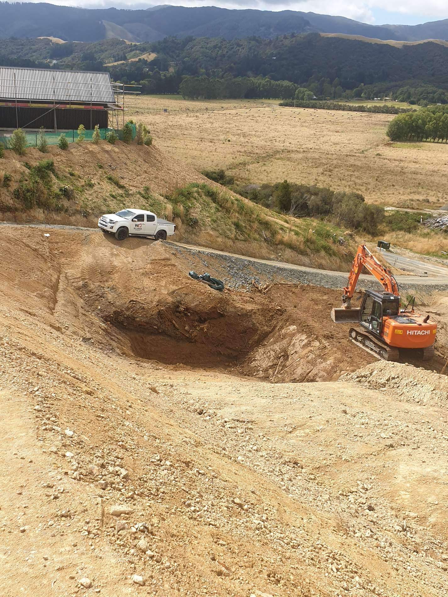 Excavator working in a large earthworks cut on a rural site, with a ute parked above and hills behind